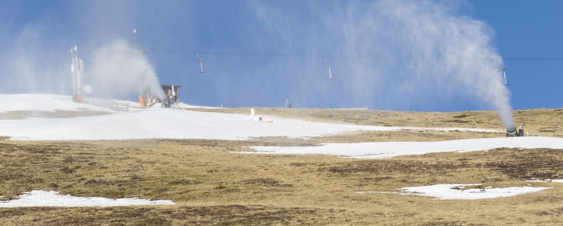 Immer mehr Flächen werden für den Alpinskisport künstlich beschneit, daneben findet sich immer öfter grüne Landschaft. (Foto: AdobeStock/mg1708) Die Kosten des Alpentourismus: Schneekanonen in Betrieb an einem nahezu schneefreien Hang im Sonnenlicht. (Foto: AdobeStock/mg1708)