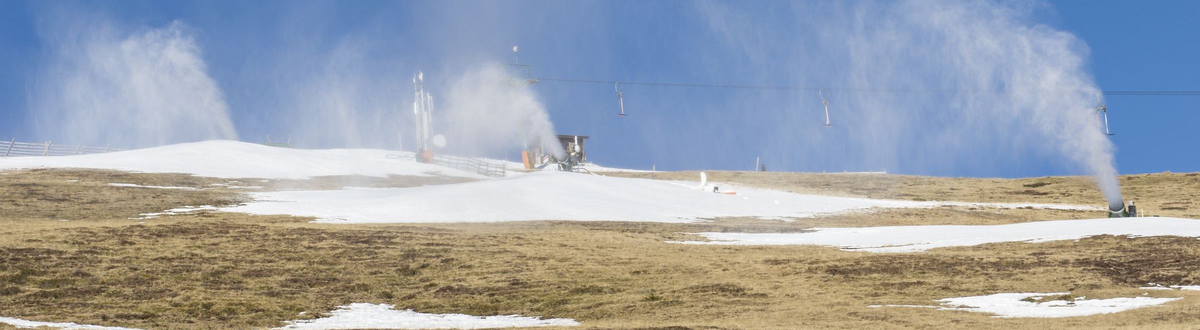 Schneekanone in den Alpen - Wintersport Schneekanonen in Betrieb an einem nahezu schneefreien Hang im Sonnenlicht. (Foto: AdobeStock/mg1708)