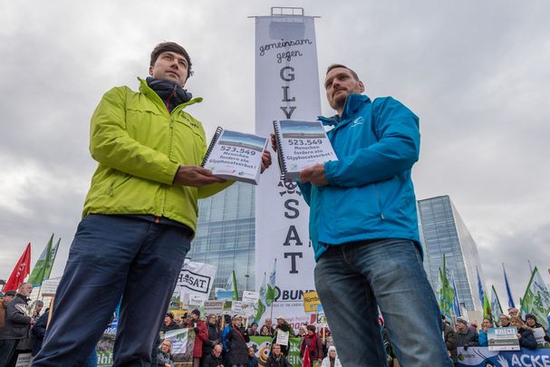 Foto: Toni Mader Zwei Männer halten Unterschriftslisten gegen Glyphosat in die Kamera, im Hintergrund sieht man Hochhäuser und Demonstranten mit Fahnen und Bannern (Foto: Toni Mader)