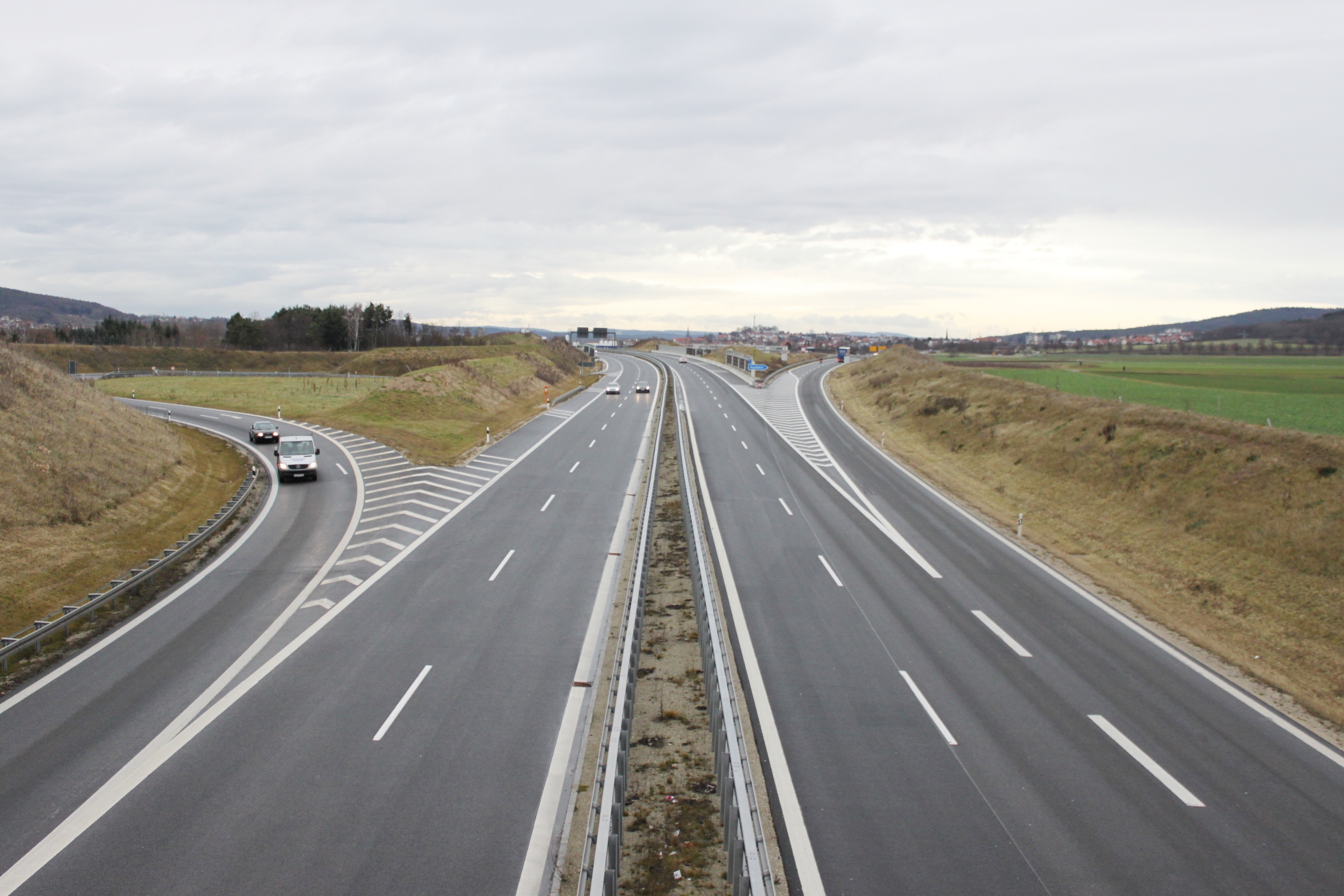 Eine mehrspurige Straße, die die gesamte Breite des Bildes einnimmt. Im Hintergrund sind Felder und ein paar Häuser zu erkennen. 