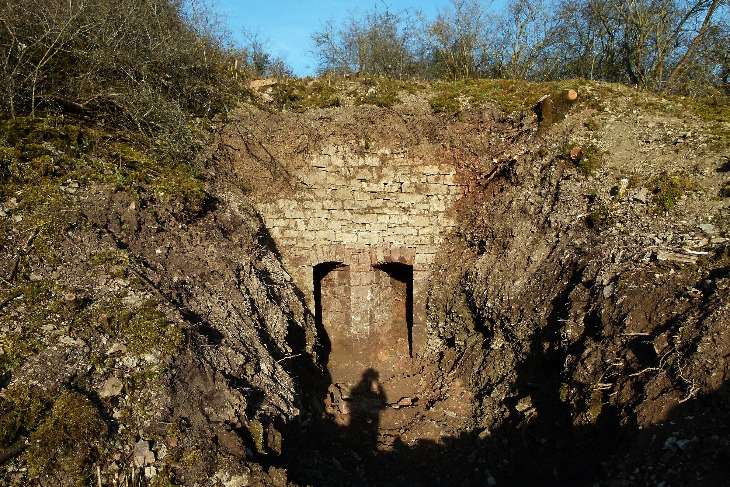 In einer freigebaggerten Grube ist im Hintergrund eine aus Steinen aufgeschichtete Mauer mit zwei bogenförmigen Öffnungen zu erkennen