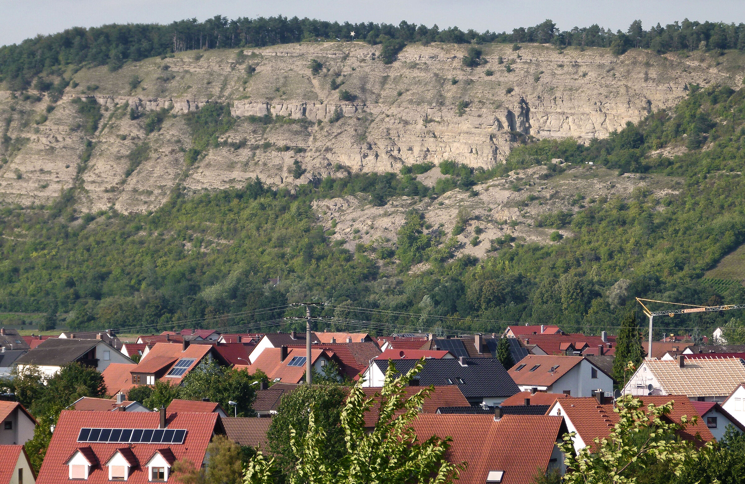 Im Hintergrund einer Ortschaft ragt eine spärlich baumdurchsetzte Felswand aufels