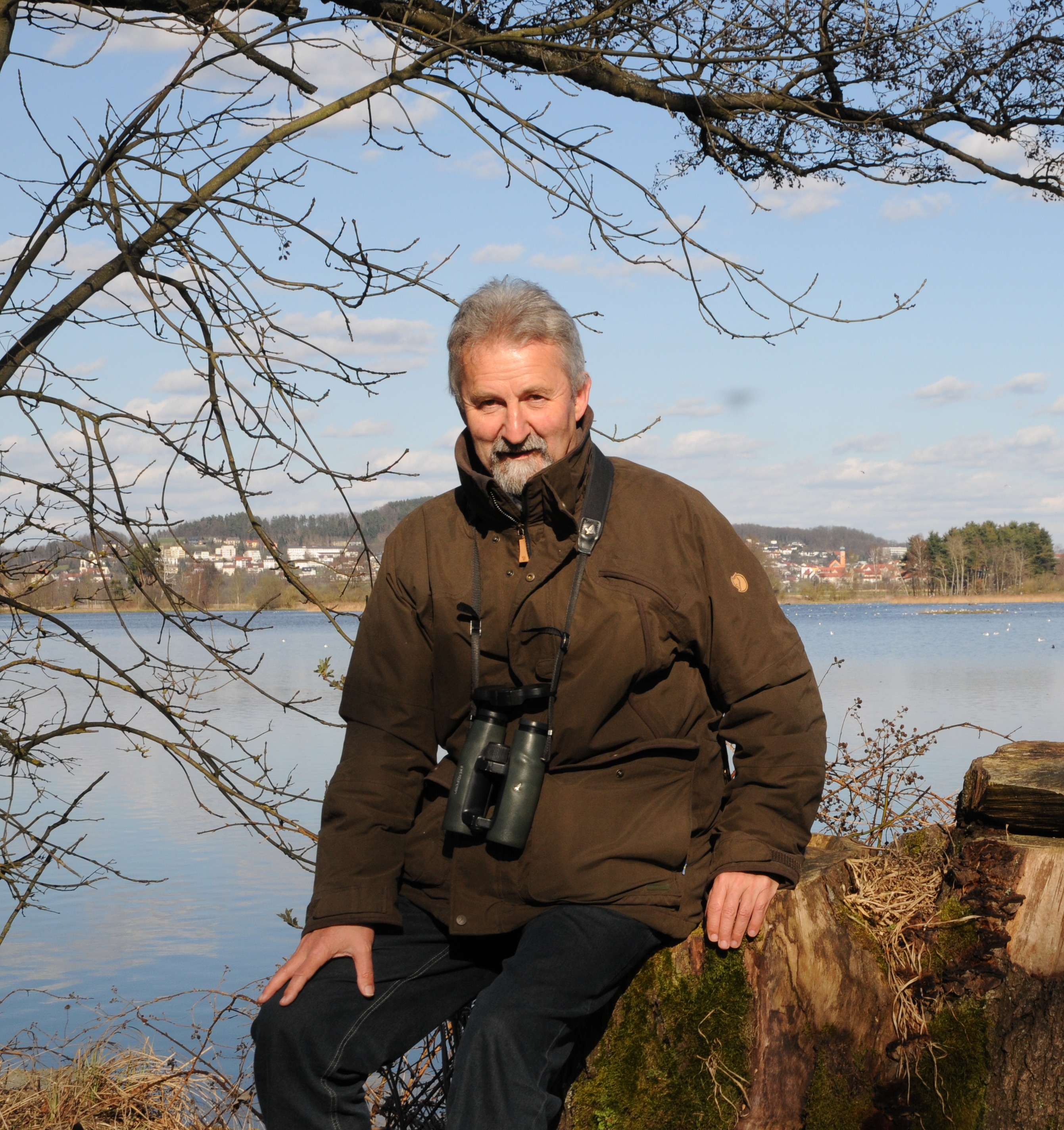 Peter Zach sitzt auf einem Baumstumpf vor dem Großen Rötelseeweiher und schaut in die Kamera. Ihm hängt ein Fernglas um den Hals. 