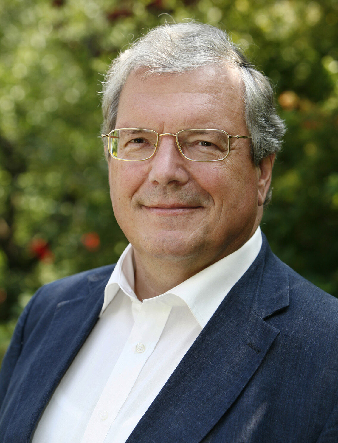 Das Portrait eines Mannes mit weißen Haaren und Brille in einem weißen Hemd und einer blauen Anzugjacke.