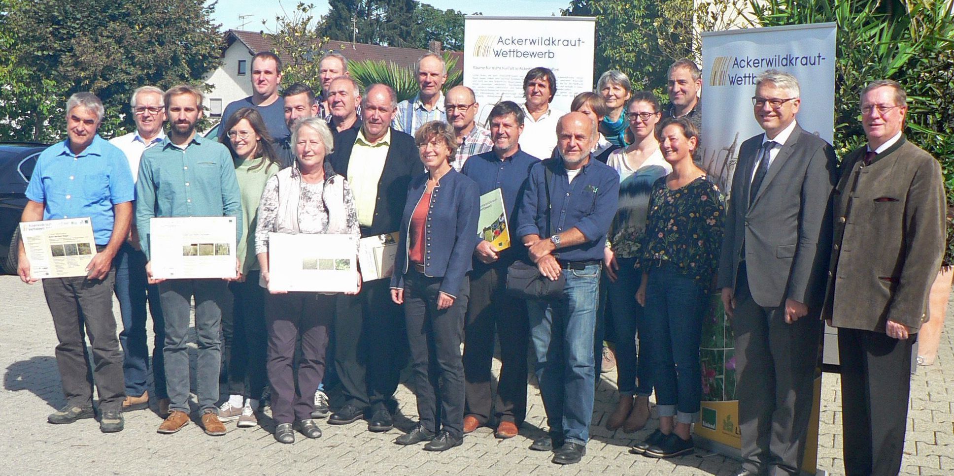Die Teilnehmer*innen des Ackerwildkraut-Wettbewerbs 2018 bei der Preisverleihung in Mamming. (Foto: LfL) Eine Gruppe Leute steht vor zwei Werbebannern für den Ackerwildkraut-Wettbewerb. Einige haben Urkunden in der Hand. Mit dem Ackerwildkraut-Wettbewerb will der BN Landwirte ermutigen, mehr Vielfalt auf ihren Äckern zuzulassen. (Foto: LfL)