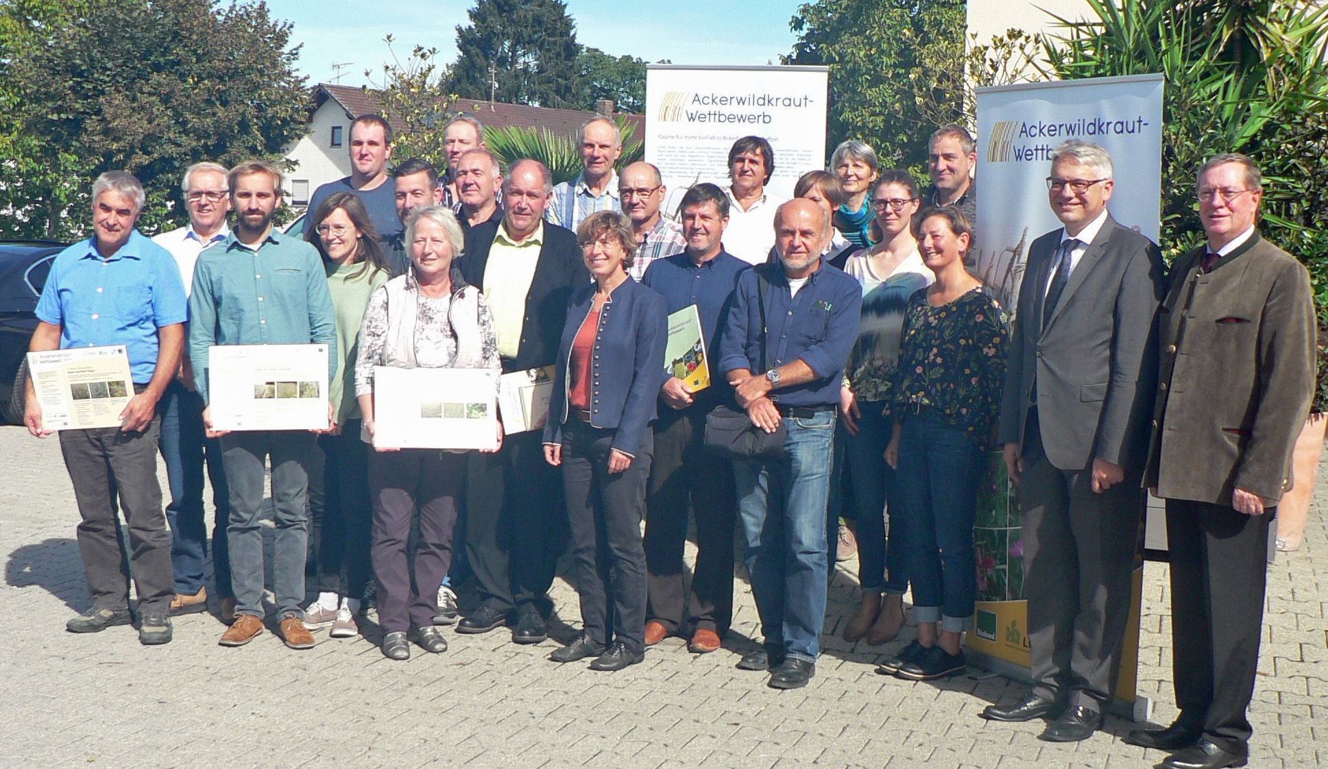 Die Teilnehmer*innen des Ackerwildkraut-Wettbewerbs 2018 bei der Preisverleihung in Mamming. (Foto: LfL) Eine Gruppe Leute steht vor zwei Werbebannern für den Ackerwildkraut-Wettbewerb. Einige haben Urkunden in der Hand. Mit dem Ackerwildkraut-Wettbewerb will der BN Landwirte ermutigen, mehr Vielfalt auf ihren Äckern zuzulassen. (Foto: LfL)