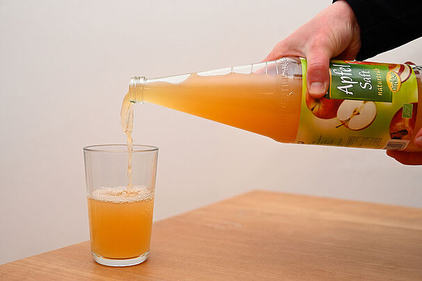 Eine Hand gießt Apfelsaft aus einer Mehrweg Flasche in ein Glas