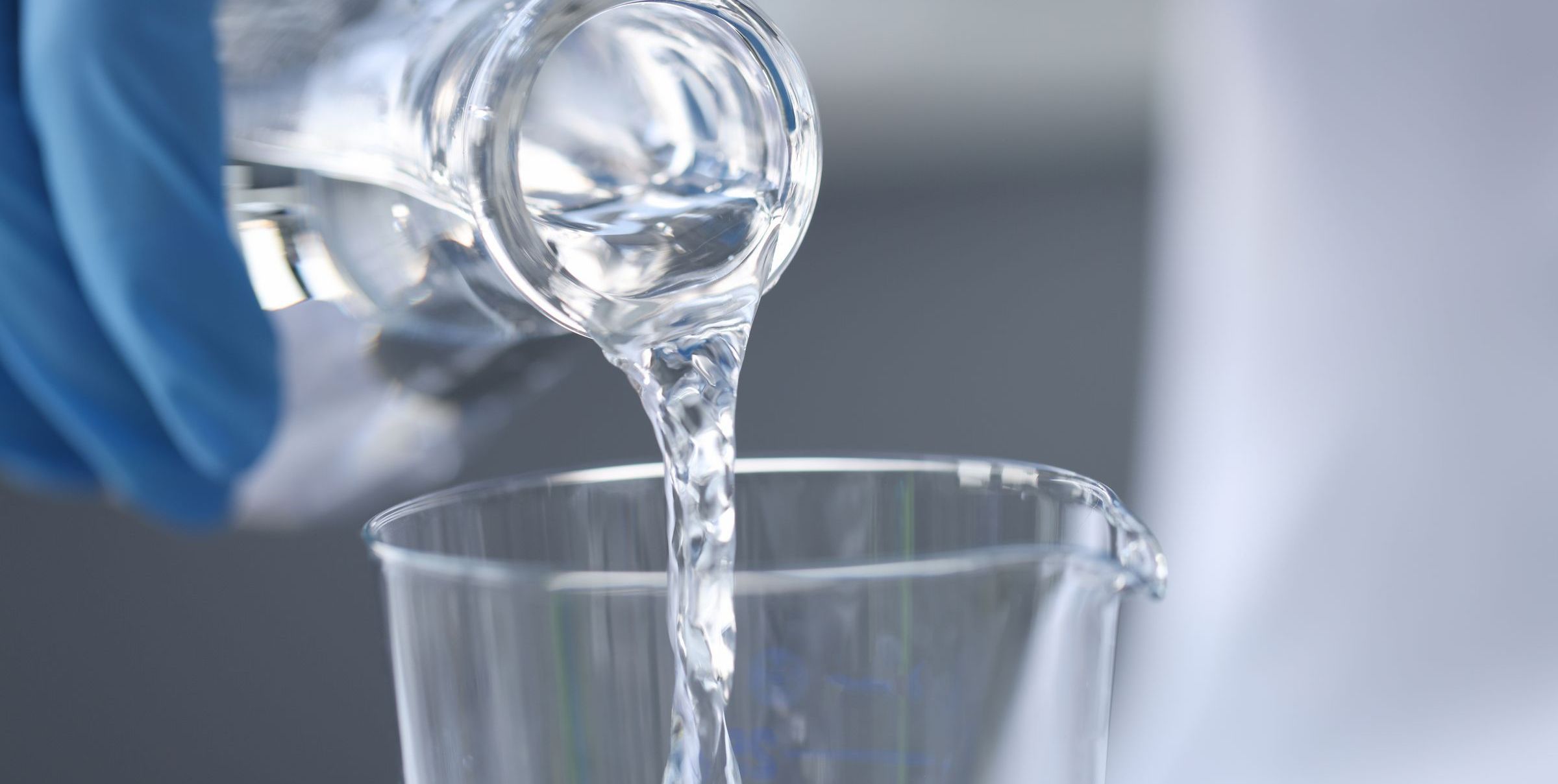 Kontrolle von Trinkwasser: Eine Hand mit Laborhandschuh gießt Wasser aus einer Flasche in ein Glas. (Foto: C_megaflopp/stock.adobe.com)