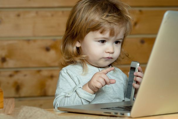 Ein Kind sitzt vor einem Laptop mit dem Smartphone in der Hand. (Foto: tverdohlib/stock.adobe.com)