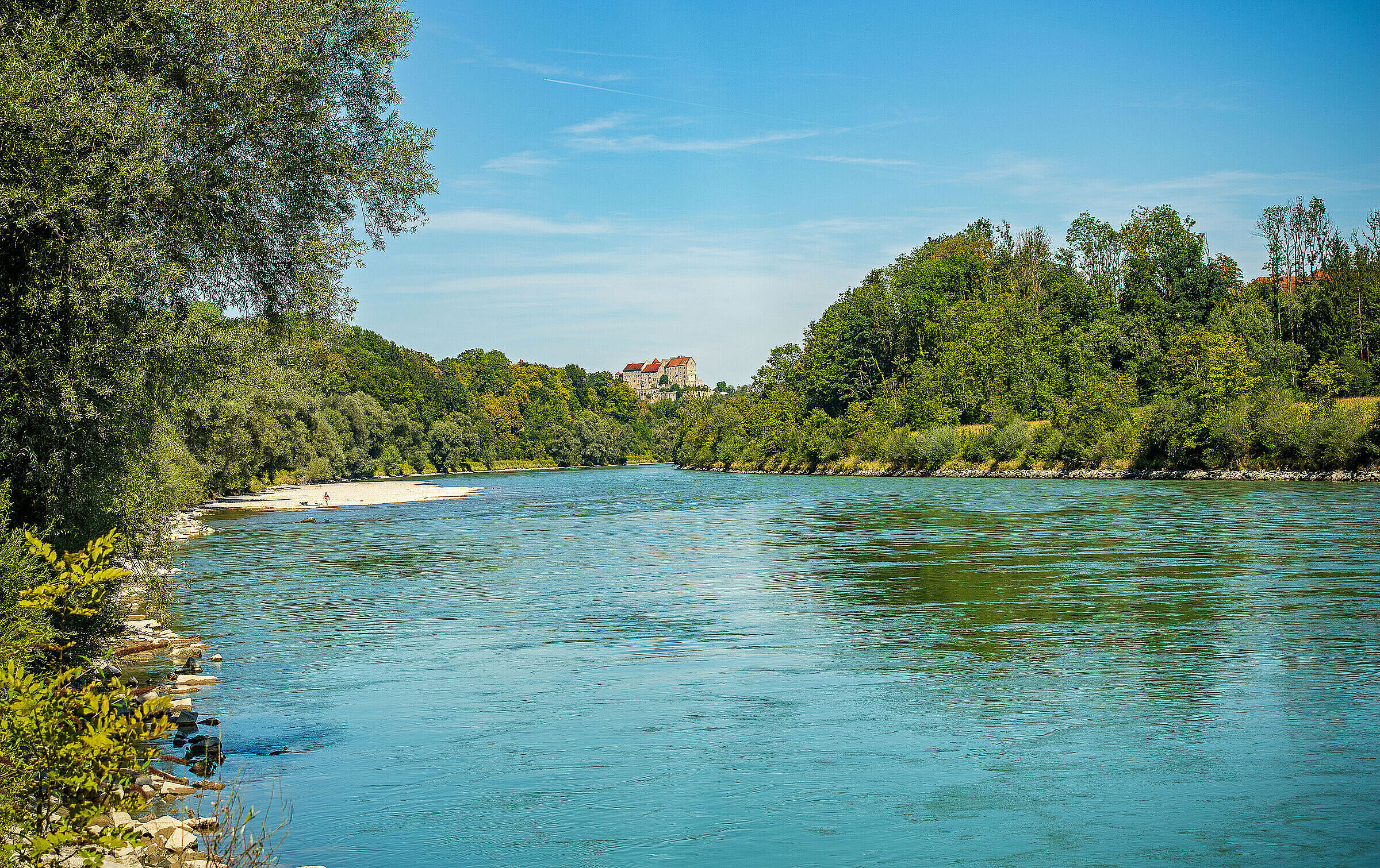 Das blaue Wasser der Salzach fließt zwischen grün bewaldeten Hügeln an Burghausen vorbei. Von der Stadt sieht man lediglich einen kleinen Teil der Burg auf einem im Hintergrund gelegenen Hügel.